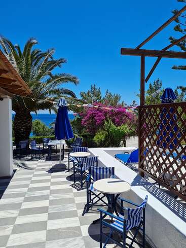 The image depicts a serene outdoor patio with a checkered floor, featuring blue and white striped chairs and tables, surrounded by palm trees and a stunning ocean view on a sunny day.