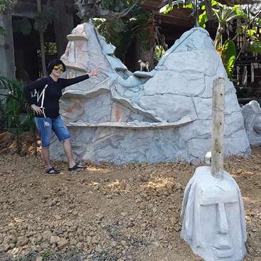 The image depicts a person posing in front of a large rock formation, with a white statue of a face in the foreground. The scene is set outdoors.