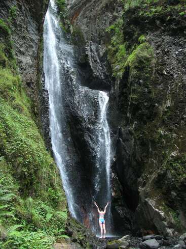 A woman stands with her arms raised at the base of a tall waterfall, surrounded by lush greenery and large rocks. She wears a blue bikini.