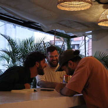 The image depicts three men seated at a bar, engaged in conversation, with a white table and ceiling, surrounded by palm trees and a cityscape visible through the windows behind them.