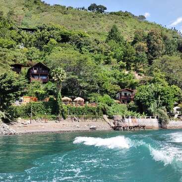 The image depicts a serene tropical landscape featuring a hillside with lush greenery, a body of water, and a small beach with a few houses and a dock.