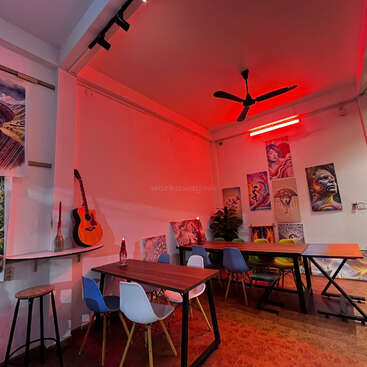 The image depicts a room with white walls, featuring a collection of paintings on the walls, a table with chairs, and a guitar on a shelf, illuminated by red lighting.