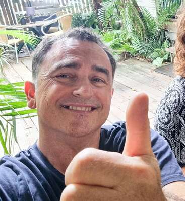 The image shows a man giving a thumbs-up gesture, with a woman\'s shoulder visible to the right and a patio area with plants and outdoor furniture in the background.
