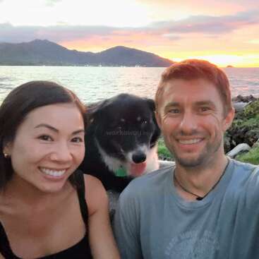 The image depicts a serene scene of a man and woman posing with their dog on a rocky beach at sunset, with mountains visible in the background.