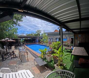 The image depicts a serene outdoor setting with a pool, surrounded by a patio area featuring lounge chairs, tables, and lush greenery, under a covered roof.