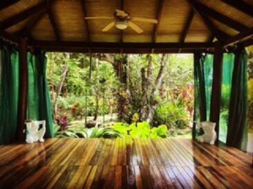 The image depicts a spacious room with a wooden floor, ceiling fan, and green curtains, featuring a serene outdoor garden view through the open windows.