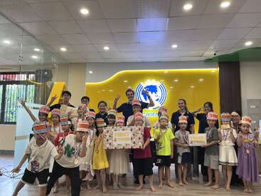 The image shows a group of children and adults posing for a photo in a classroom, holding signs and wearing medals, with a yellow wall featuring a logo behind them.