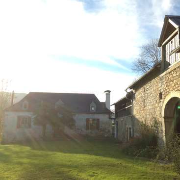 The image depicts a serene scene of two houses with stone exteriors, one with a chimney, set amidst lush green grass and a blue sky with wispy clouds.