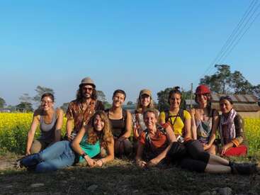 The image depicts a group of nine people sitting on the ground in a field of yellow flowers, posing for a photo on a sunny day.