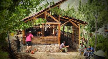 The image depicts a group of people working together to build a structure, likely a house or building, with a focus on teamwork and collaboration. The scene is set in a rural area.