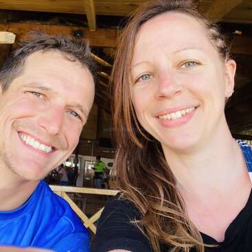 The image shows a selfie of a man and woman with fair skin, smiling at the camera, in front of a wooden structure with people in the background.