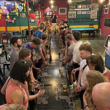 The image depicts a lively gathering of people in a bar, standing around a long table laden with drinks, with a pool table and dartboard visible in the background.