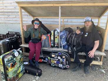 The image depicts a man and woman sitting on a wooden bench, surrounded by luggage and musical equipment, with a rustic log cabin wall in the background.