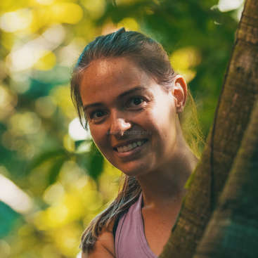 The image features a woman with brown hair, wearing a purple tank top, standing in front of a tree trunk, with a blurred background of greenery and yellow leaves.