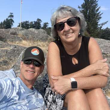 The image depicts a man and woman sitting on rocks, with the man wearing a hat and the woman wearing sunglasses, set against a backdrop of trees and a blue sky.