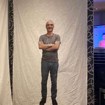 The image depicts a man standing in front of a white backdrop, with his arms crossed, wearing a gray t-shirt and jeans, in a studio-like setting.