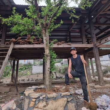 A man in a black tank top and cap stands beside a tree in front of a wooden structure with a corrugated metal roof, holding a shovel.