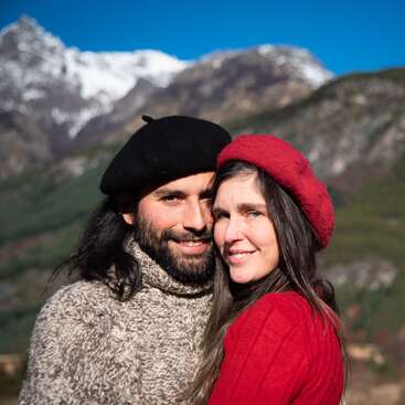 The image depicts a man and woman embracing in front of a mountainous backdrop, with the woman wearing a red hat and the man sporting a black beret.