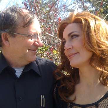The image depicts a man and woman standing together, facing each other, with the woman\'s face blurred out. The man is wearing a dark shirt and has short hair.