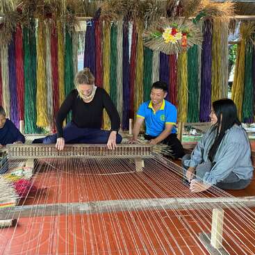 The image depicts a group of people engaged in traditional weaving, surrounded by vibrant threads and colorful decorations, in a serene outdoor setting.