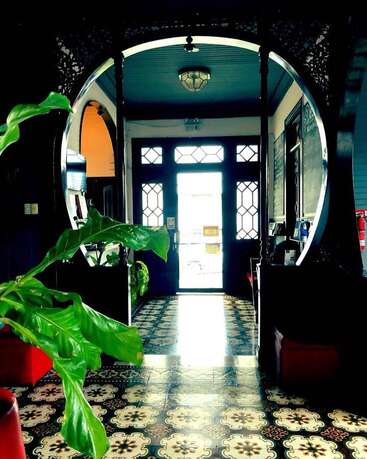 The image depicts a hallway with a large arched doorway, patterned tile floor, and a plant in the foreground, bathed in natural light.