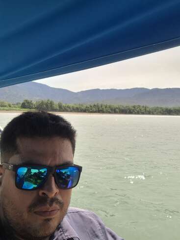 The image depicts a man on a boat, with a serene body of water and a mountainous landscape in the background, set against a bright and sunny day.