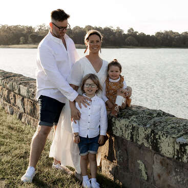 The image depicts a family of four posing for a photo by a stone wall overlooking a serene body of water on a sunny day.