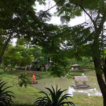 The image depicts a serene pond scene with a small hut on the water, surrounded by lush greenery and trees, set against an overcast sky.
