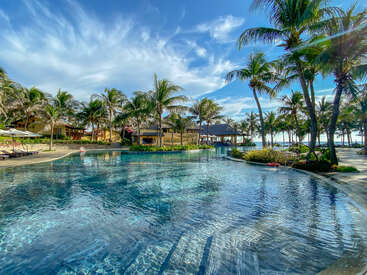 The image depicts a serene pool scene with palm trees, a blue sky, and a building in the background, exuding a tropical and relaxing atmosphere.