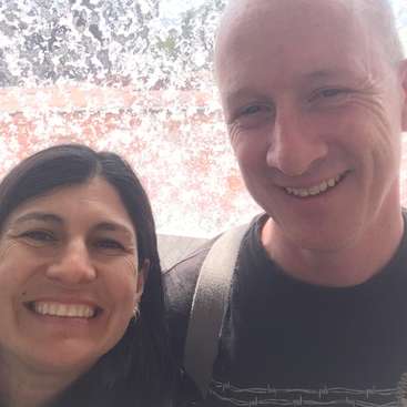 The image shows a selfie of a man and woman smiling at the camera, with the woman on the left and the man on the right, both wearing dark clothing.