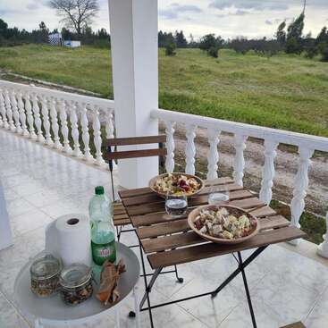 The image depicts a serene outdoor setting with a table and chairs on a porch, featuring a bowl of salad, a glass of water, and a bottle of juice, set against a picturesque grassy field.
