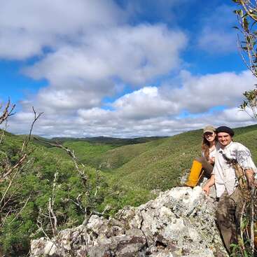 The image depicts a serene mountainous landscape with two individuals standing on a rocky outcrop, surrounded by lush greenery and a blue sky with white clouds.