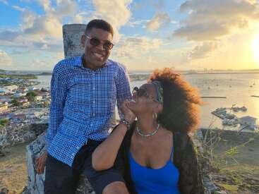 The image depicts a man and woman sitting on a rocky outcrop overlooking a coastal town, with a body of water and boats in the background at sunset.