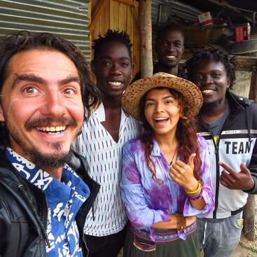 The image shows a group of five people posing for a selfie in front of a corrugated metal wall, with a wooden structure behind them.