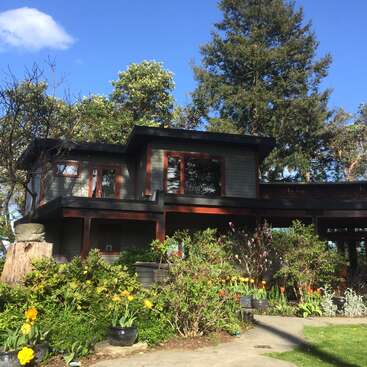 The image depicts a two-story house with a dark exterior, surrounded by lush greenery and vibrant flowers, set against a serene blue sky.
