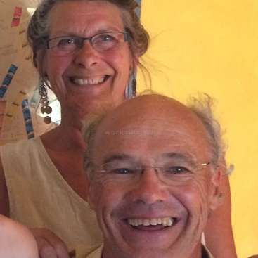 The image depicts a close-up of a man and woman smiling, with the woman behind the man, set against a yellow wall.