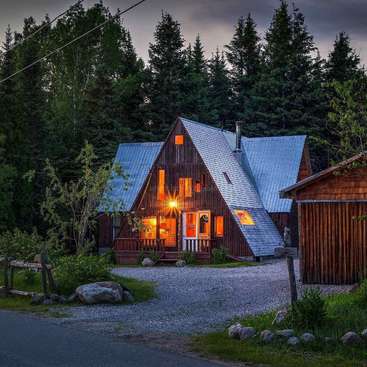 The image depicts a charming A-frame house with a lighted front door, nestled among trees, featuring a gravel driveway and a wooden fence, exuding a cozy and inviting atmosphere.