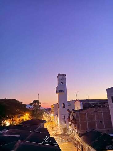The image depicts a serene cityscape at dusk, featuring a prominent white clock tower, buildings, and a vibrant sky with hues of purple, pink, and orange.