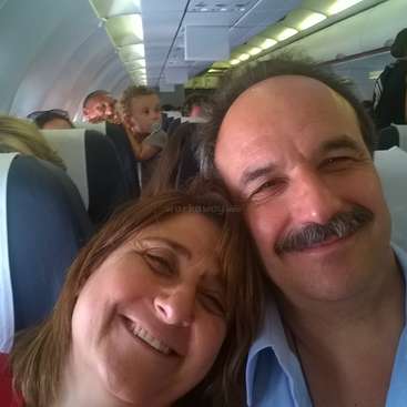 The image shows a selfie of a man and woman on an airplane, with other passengers in the background, likely taken during a flight.