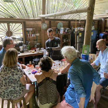 The image depicts a group of people gathered around a table in a rustic kitchen, with a man standing at the center, surrounded by various kitchen items and utensils.