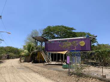 The image depicts a purple shipping container house with a yellow sun logo, situated on a dirt road, surrounded by trees and a clear blue sky.