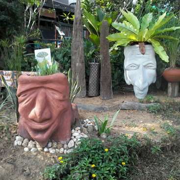 The image depicts a serene garden scene featuring two large, intricately designed planters shaped like human faces, surrounded by lush greenery and vibrant flowers.